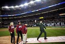 A man in a green military uniform prepares to throw a ball, while two women and a man on the left wearing red jackets with "World Series" on the chest look on.