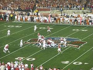 Several American football players in red and white uniforms  in action at the mid-field area of the stadium with a large logo visible on the field. Players are visible on both sidelines with the edge of the spectator stands also visible.