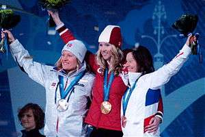 Three smiling women stand side-by-side with one arm over each other's back. Each holds high a flower bouquet and wears a medal around the neck. On the left, a long-haired blond wears a shiny light-gray jacket and winter cap with a badge bearing the Norwegian flag and Olympic rings. On the center, another long-haired blond wears a red jacket and red-and-white winter cap. On the right, a long-haired brunette wears a white jacket with blue and red stripes on the back.