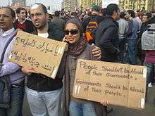 A young man and a woman carrying the tops of cardboard boxes in a crowded group of people. On one boxtop is written "People shouldn't be afraid of their governments. Governments should be afraid of their people" in English and on the other, two questions in Arabic. He has a bandage on his forehead and she is wearing a headscarf and sunglasses.