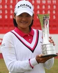 A woman in all white clothing with a red polo golf shirt underneath the sweater and the wrist and neck seems are colored in magenta and blue with a white golf hat with black lettering and a silver metal trophy in hand