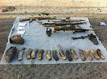 Various weapons, including assault rifles and ammunition canisters, laid out on a blanket on the ground