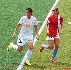 Wambach in a white uniform surrounded by a defender in a red uniform.