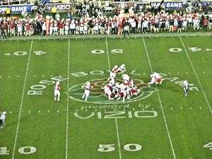 Several American football players in red and white uniforms in action at the mid-field area of the stadium with a large logo visible on the field.
