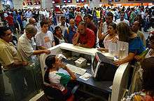 Busy interior of an airport terminal, with people crowding around a ticket counter, where a woman ticket agent is seated, listening patiently to an animated customer waving her hand.