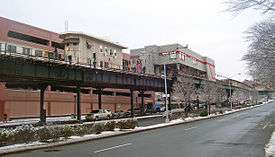 An elevated railroad track above a city street in winter, with two buildings. The smaller one, on the left, is pale yellow and narrow. The larger one at right, gray with a red stripe, is box-shaped and elevated over the tracks