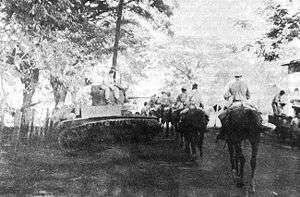 A long column of men on horseback moving down a road. A tank is parked beside the road