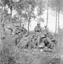 A group of soldiers in a field, some standing and some siting, looking at maps and other documents on the ground, while in the background is a stand of trees