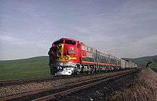 Red diesel locomotive with yellow striping leading silver train cars through rolling countryside