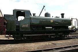 A pannier tank locomotive is standing outside an engine shed. The locomotive is mainly painted green above the running plate, although the chimney is black. On the side of the pannier tank a yellow circle encloses the yellow letters "G W R". The G and R are shaped to match the curve of the enclosing circle.
