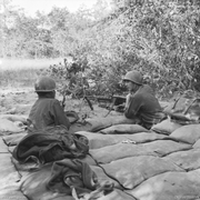 Two soldiers manning a machine-gun in a heavily sandbagged pit observe the perimetre of their defensive position through the vegetation.