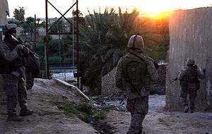 Three Marines patrolling through an Iraqi town near a river as the sun sets.