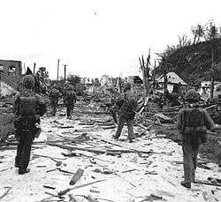 A group of Marines in a tactical column patrolling down a street through the ruins of a devastated town.