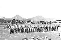 A group of Marines standing at attention on a basketball court, with Hawaiian mountains in the background.