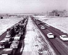 A black-and-white photo shows a four-lane freeway divided by a grass median. In the oncoming lanes, traffic is congested into the distance. With few exceptions, the freeway is surrounded by farmland.