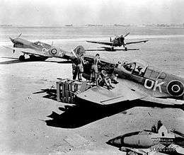 Several men in shorts working on a single-engined fighter plane; two other fighters are parked a short distance away