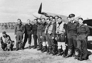 Thirteen men wearing military uniform standing close together in front of a single propeller monoplane. A fourteenth man wearing military uniform is crouching and patting a dog to the left of the group.