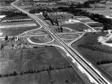 A black-and-white photo of a cloverleaf interchange between a freeway and surface road surrounded by farmland and trees