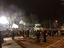 People mill about a fire and larger gatherings at night on a wintry street by a police building
