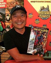 A photograph of a man wearing a black baseball cap and a black t-shirt looking at the viewer and smiling while holding a signed comic book