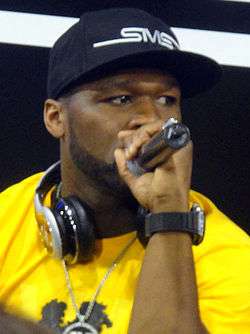 An African-American man, wearing a yellow shirt and baseball cap, speaks into a microphone.