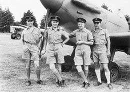 Four men wearing military uniforms posing in front of a propeller-driven aircraft