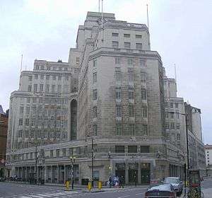 Two wings of a large white stone office building with regularly spaced rectangular windows. The building rises to twelve storeys, stepping back to a central tower surmounted by a clock and a flagpole.