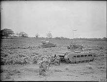 In the foreground, a group of soldiers crouch behind a tank. Another group, barely visible, crouch behind another tank in the background.