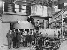 Six men in suits and ties stand in front of gigantic machinery. Two more are sitting in top of it.