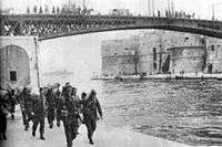 Soldiers carrying kit-bags approaching the photographer, while walking on an embankment next to a river with steel girder bridge in the background.