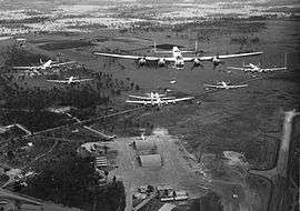 Head-on view of seven four-engined military aircraft flying over an airfield