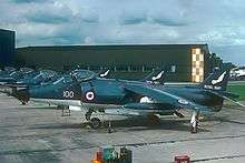 Line-up of Sea Harrier jet aircraft, facing left of photograph. In the distance is a tall, dull-coloured warehouse.