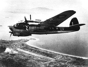 Twin-engined military aircraft in flight high over a seashore