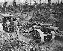 A large field gun and limber being towed along a sunken road through a battle-scarred forest by a Holt tractor. The gun has very large fabricated steel spoked wheels, with a wide tread to cope with mud.