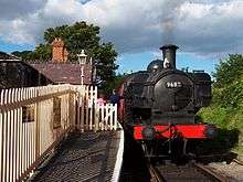 A black pannier tank locomotive is standing at the platform of a small rural railway station with a brick and slate building, cream wooden fencing, and lamp. The driver is standing next to the locomotive and is talking to a group of passengers who are about to board the red passenger carriage.