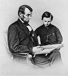 A seated Lincoln holding a book as his young son looks at it