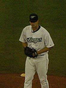 A man in a white baseball uniform reading "Jays" across the chest, dark baseball cap, and black baseball glove stands on a dirt mound in a grass field.