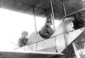 Two men in flying gear seated in tandem open cockpits of a biplane