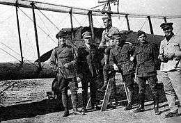 Full-length outdoor portrait of six men in military uniforms in front of a military biplane with a machine gun mounted on the upper wing