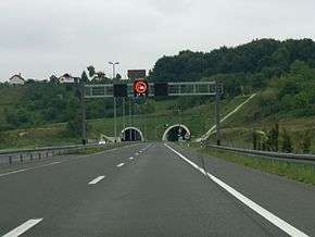 Variable traffic signs placed on a gantry spanning three motorway lanes. The sign informs of overtaking ban for freight vehicles being enforced and that current temperature is 17 degrees Celsius. A two-tube tunnel portal is visible in the background.
