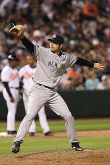 A man in a grey baseball uniform with "New York" on the chest wearing a baseball glove and a dark cap winds up to throw with his left hand.