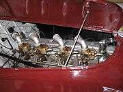 Close-up of a straight-eight engine installed in the front of a racing car, as seen through the right side of the bonnet, which is hinged in the centre and is held open with a metal rod. There are four downdraft carburettors on the near side of the engine, these have intake stacks angled backward and toward the engine.  The exhaust manifold is visible on the same side of the engine as the carburettors.