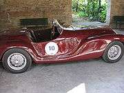 Right side view of a sports car shaded by a brick wall. The number "10" is written inside a circle on the driver's door. The car has wire wheels, drum brakes, bucket seats, a trunk, a wooden steering wheel, a racing windshield, and no top.