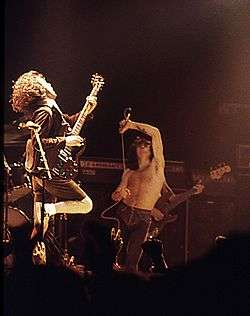 AC/DC guitarist Angus Young (in requisite school blazer, hat and shorts) and lead singer Bon Scott (in black trousers and topless) in an energetic performance on stage.