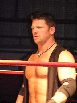An adult white male wearing a black vest standing in a wrestling ring with red ropes.
