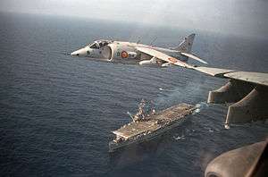 A Harrier flies over an aircraft carrier below