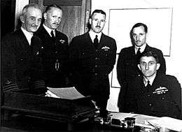 Five men, four of whom are standing and one seated, wearing dark-coloured military uniforms