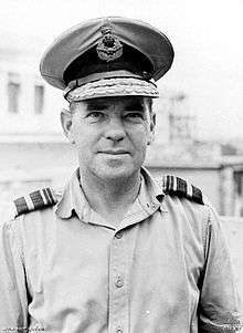 Outdoor head-and-shoulders portrait of man in light-coloured shirt with shoulder insignia, wearing peaked cap with two rows of braid
