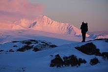 A hiker standing on a rocky knoll, the pink early morning light illuminating snow-covered Mt. Moffett in the background