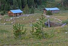 Two log cabins with metal roofs in the distance down a rolling slope. The remains of another are partially visible closer to the camera